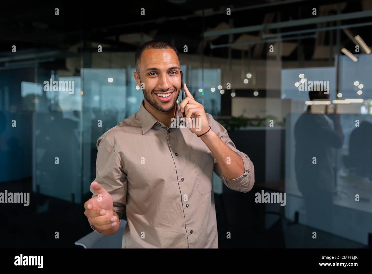 A young African American man is standing in the office and talking on the phone. gesturing with his hands, looking at the camera, smiling. Stock Photo