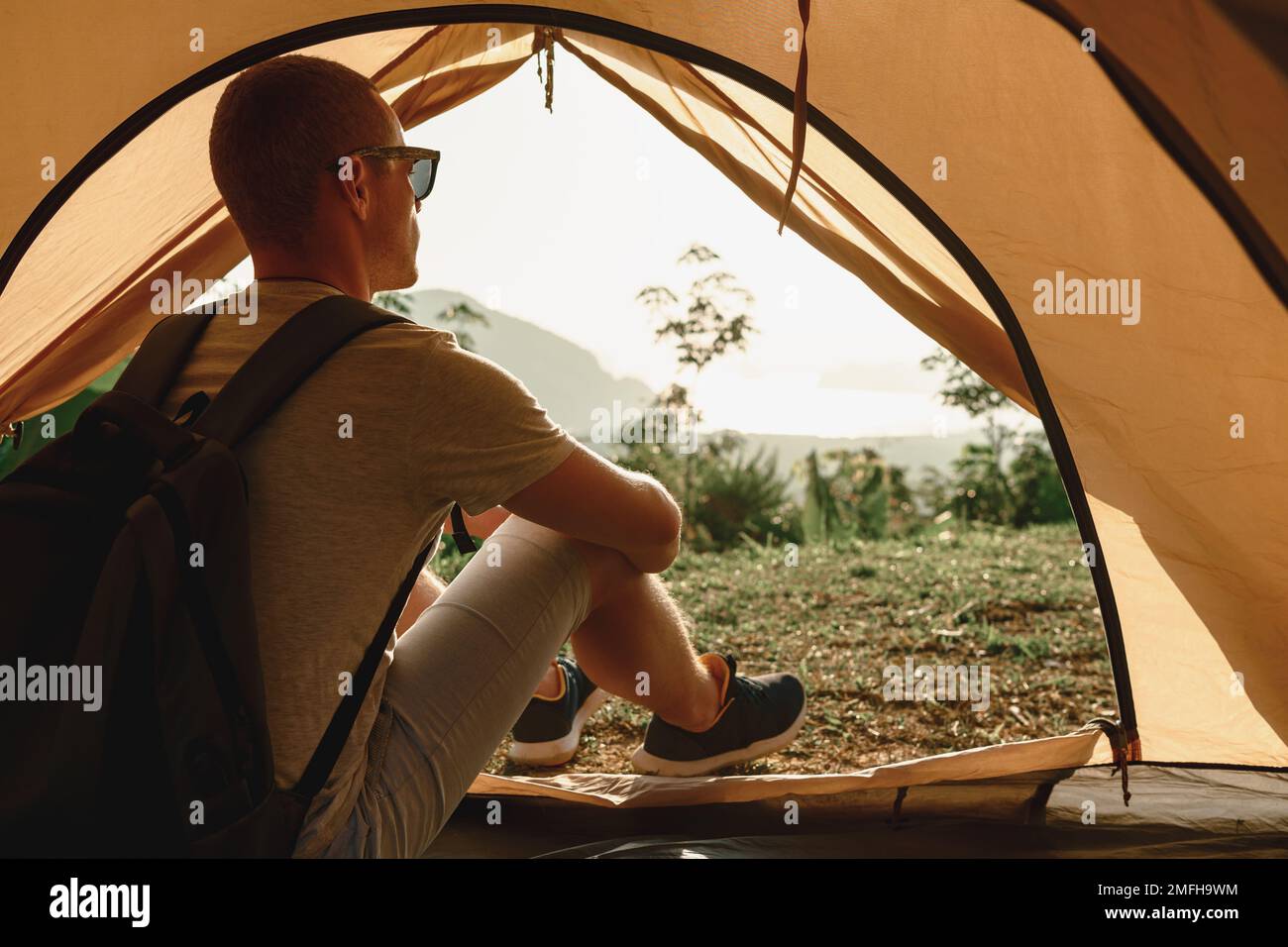 One young hiker man with backpack sits in tent and looks at sunrise or sunset Stock Photo