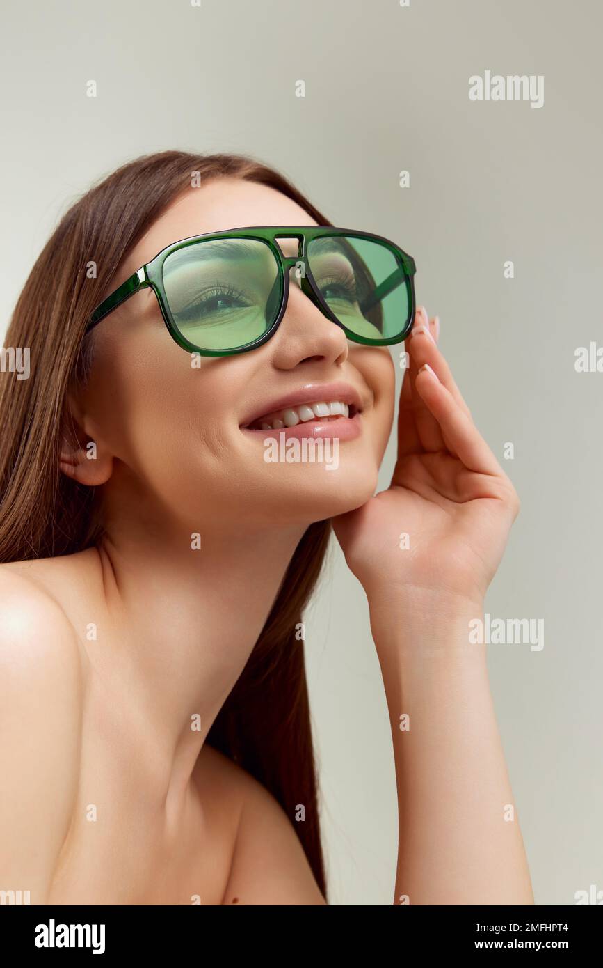 Portrait of young beautiful girl in green sunglasses posing, smiling over grey studio background. Perfect smile. Concept of beauty, fashion Stock Photo
