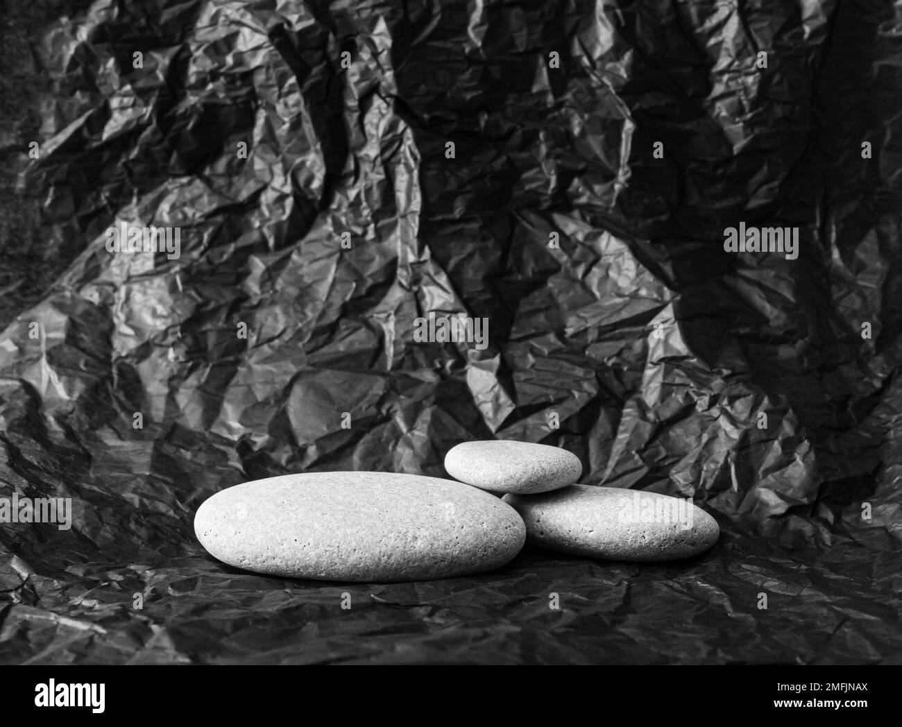 Gray color flat sea stones stacked on black color crumpled abstract silk paper products advertising background, side studio light. Lot of copy space. Stock Photo