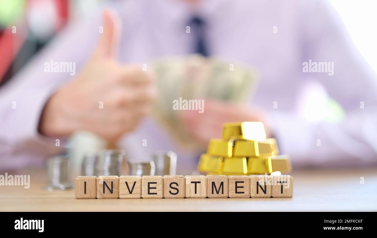 Word investment on cubes, stacks of coins and gold bars and investor with thumbs up in background. Stock Photo