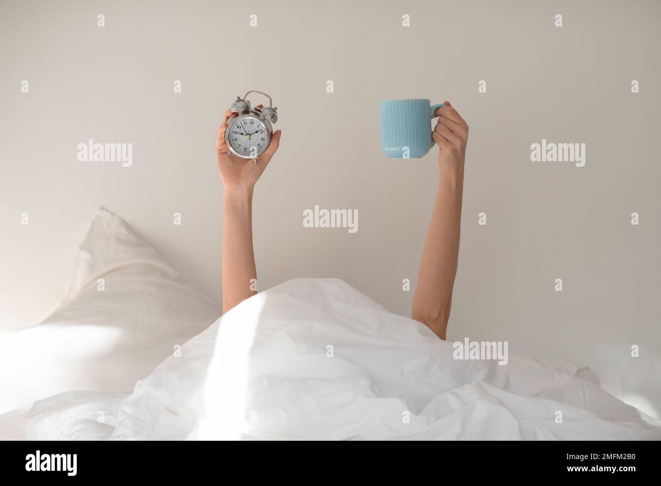 Hands of young european woman under covers with alarm clock, cup of coffee on white bed in bedroom Stock Photo
