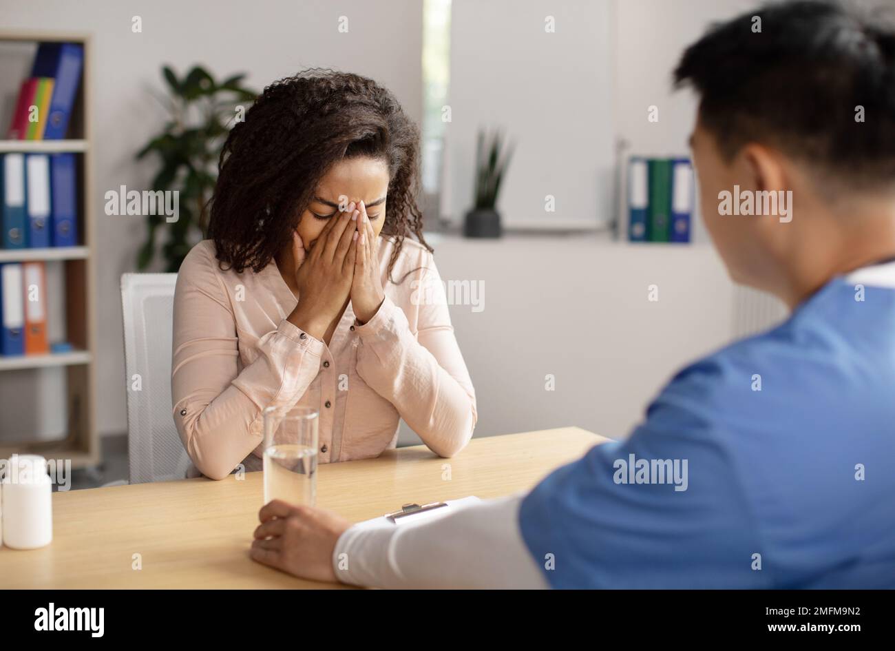 Serious middle aged korean doctor calms crying black millennial female patient, giving glass of water Stock Photo