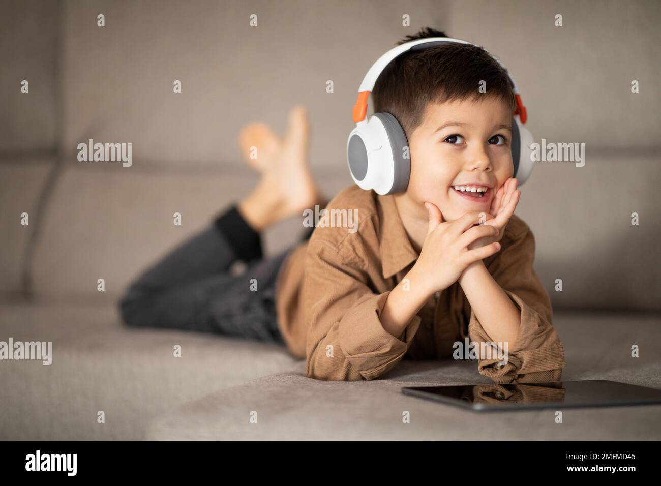 Happy pensive little boy in wireless headphones thinks, creative idea, watching video on tablet, lies on sofa Stock Photo