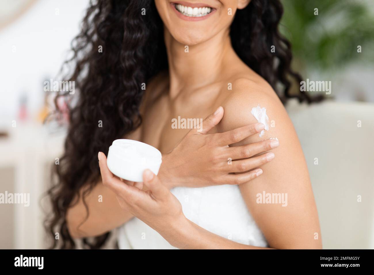 Cheerful islamic millennial curly lady in towel holding jar with copy space, apply cream on shoulder, enjoy procedure Stock Photo