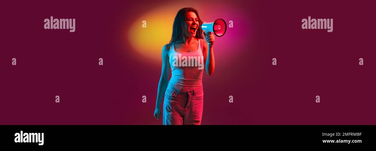 Emotive young girl in casual clothes shouting in megaphone over gradient dark pink background in neon light. Information, news. Banner, flyer Stock Photo