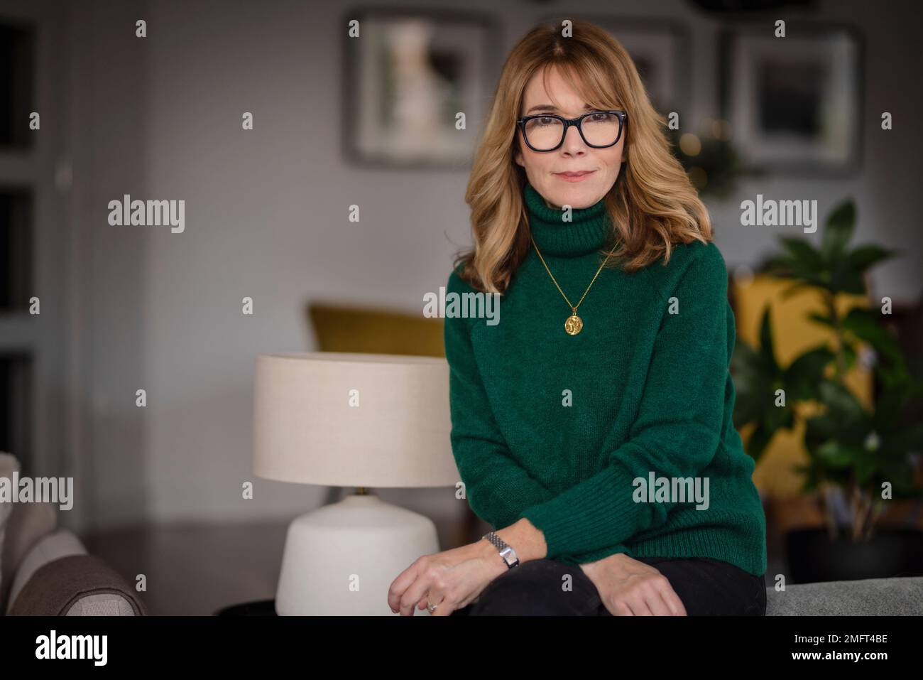 Portrait of attractive middle aged woman relaxing in an armchair at home. Blond haired female wearing eyeglasses and sweater. Stock Photo