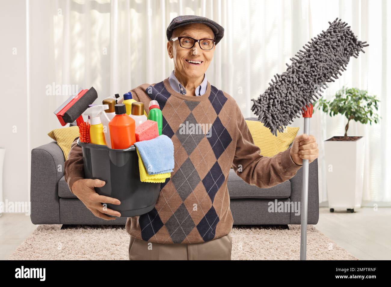 Elderly man holding a bucket with cleaning supplies and a floor mop in a living room Stock Photo