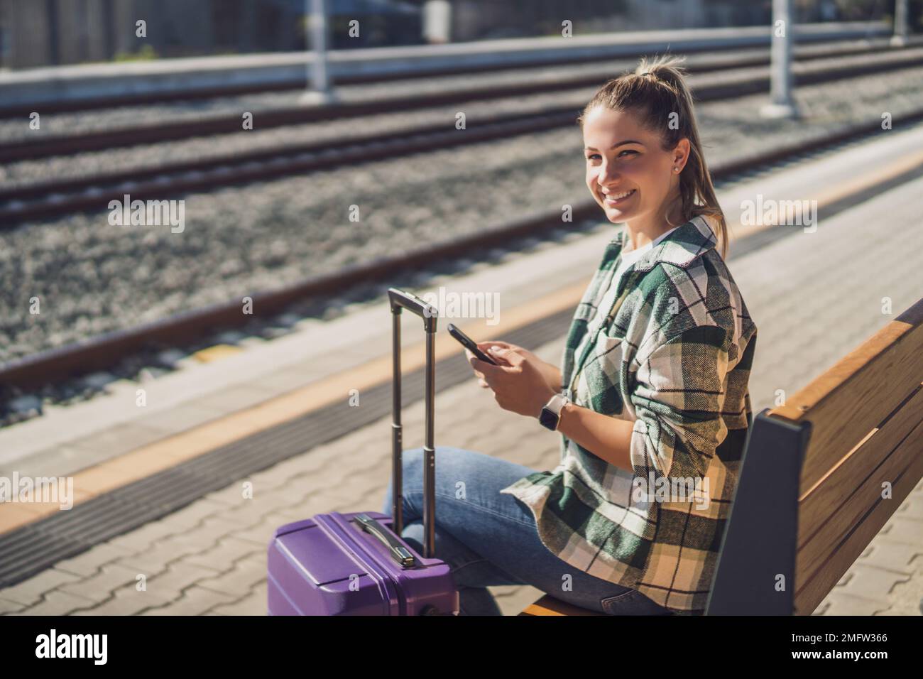 Happy woman using mobile phone while sitting on a bench at the train station. Stock Photo