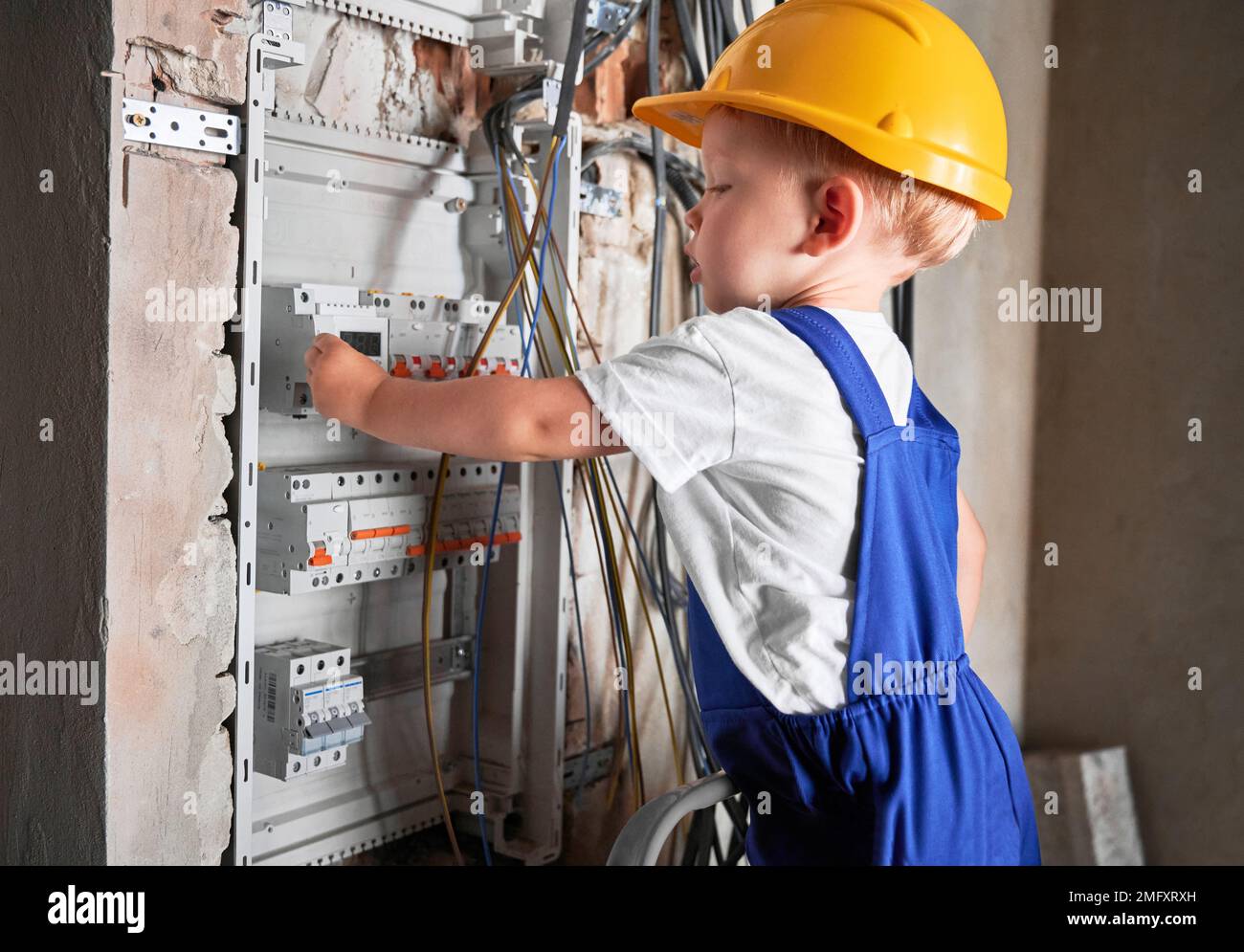 Side view of child in safety helmet fixing electrical switchboard or distribution board in apartment under renovation. Baby electrician repairing household electrical panel with automatic fuses. Stock Photo