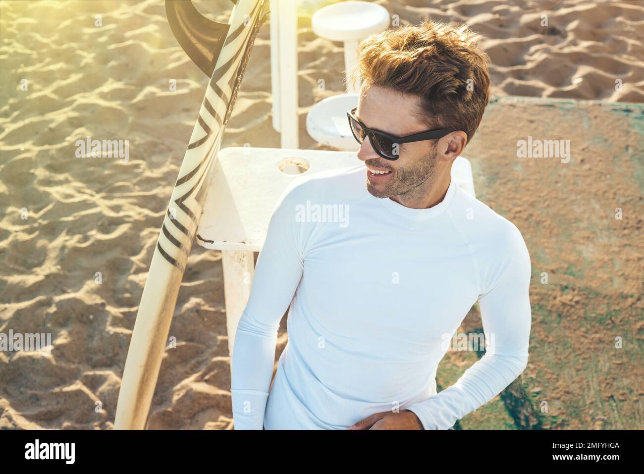 Young handsome smiling man is sitting on a beach next to his surfboard ...