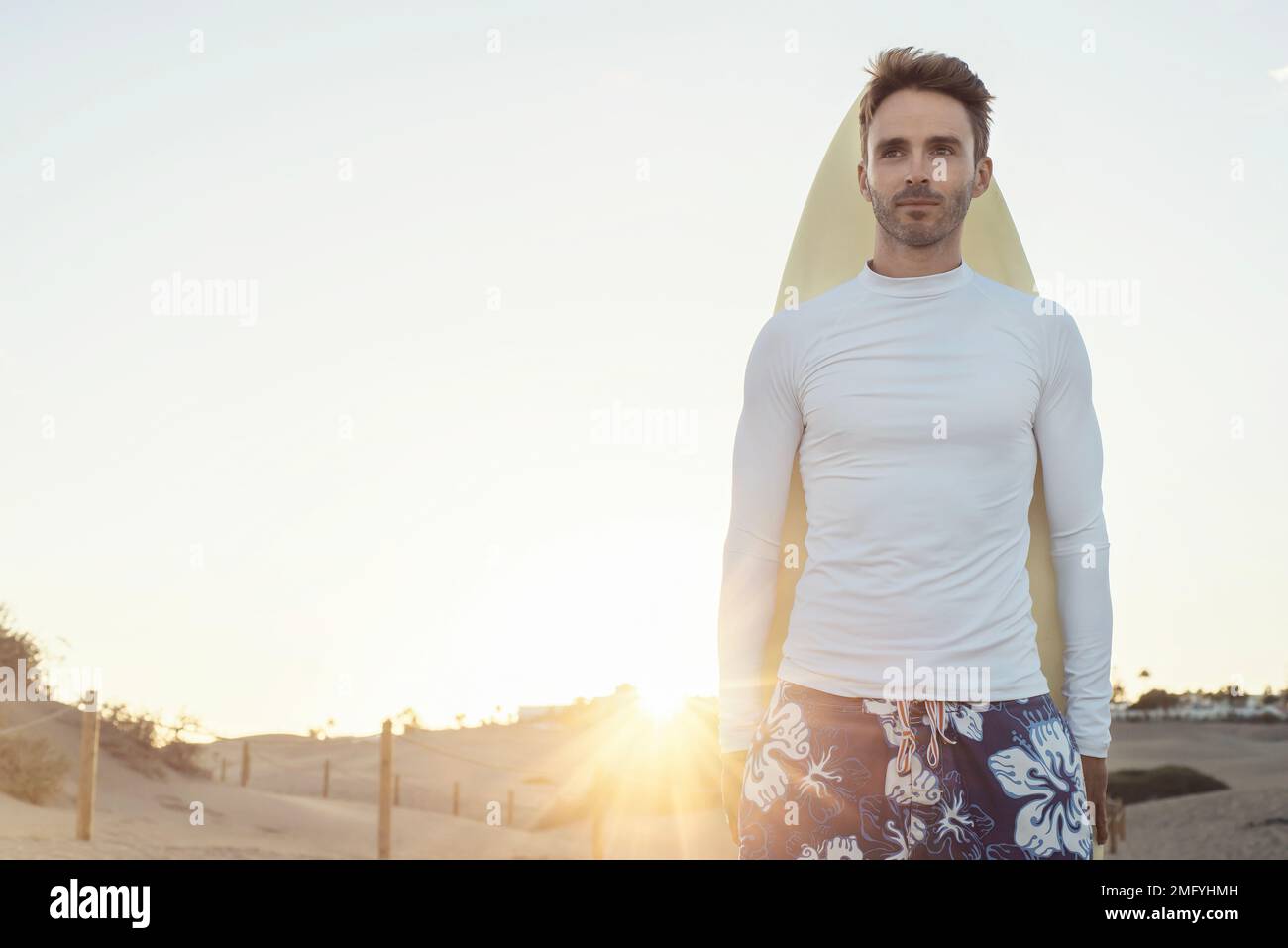 Young handsome surfer guy is standing next to his surfboard and wearing white, empty, blank lycra shirt. Horizontal mock up with sun light flare. Stock Photo