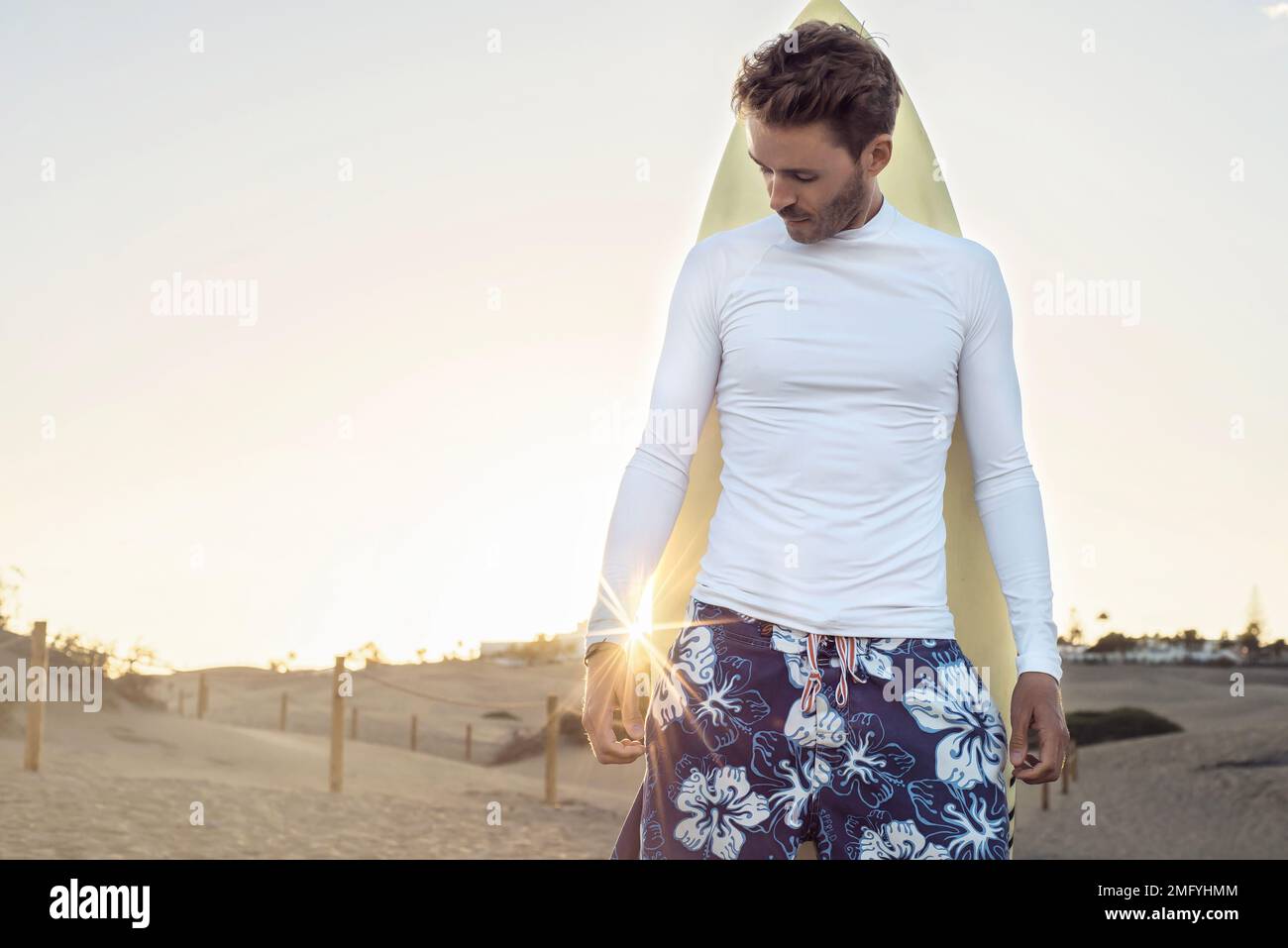 Young handsome surfer guy is standing next to his surfboard and wearing white, empty, blank lycra shirt. Horizontal mock up with sun light flare. Stock Photo