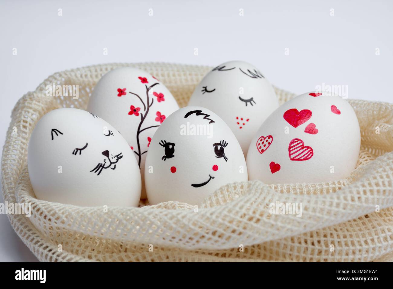 Close-up Delicately painted Easter eggs lie in environmentally friendly mesh bag on white table. Funny peaks and twig with red flowers are drawn on eggs. Copy space. Happy Easter Stock Photo