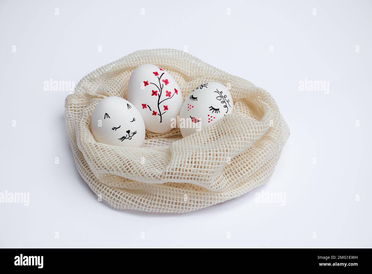 Three beautifully painted Easter eggs lie in environmentally friendly mesh bag on white table. Funny faces and twig with red flowers are painted on eggs. Copy space. Happy holidays Stock Photo