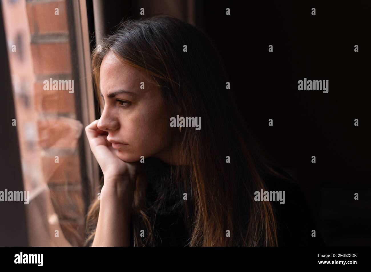 Side view of young thoughtful female with long dark hair leaning chin on hand and looking out window at home Stock Photo