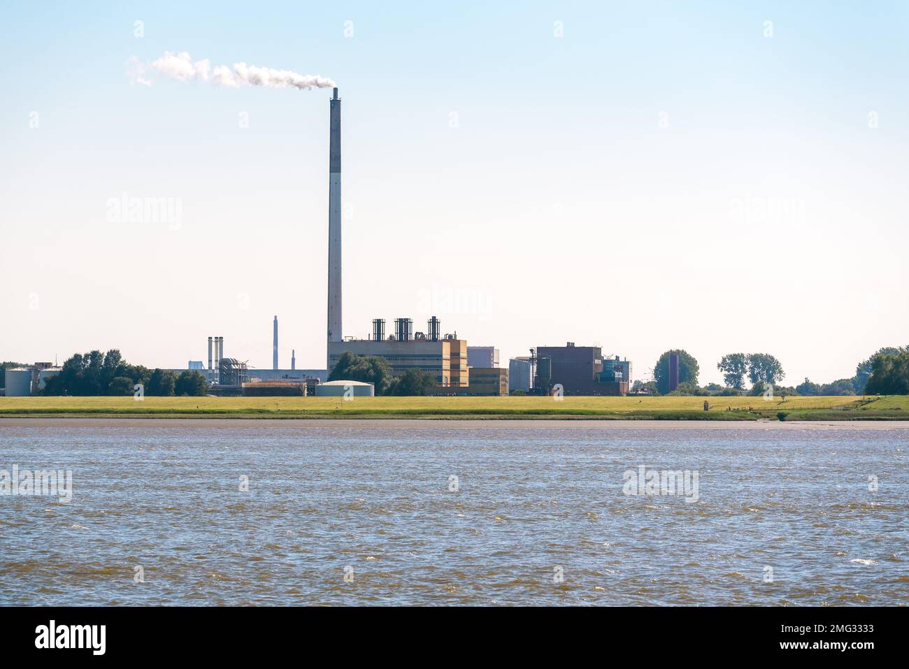 Chemical plant with a tall smokestack belching out white smoke on the bank of a river on a clear summer day Stock Photo