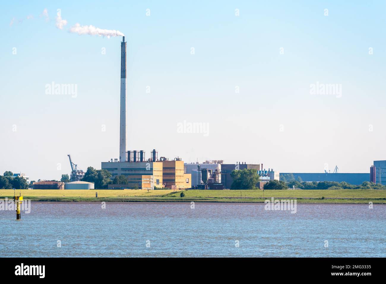 View of a riverside chemical factory with a tall smokestack belching out white smoke at sunset Stock Photo