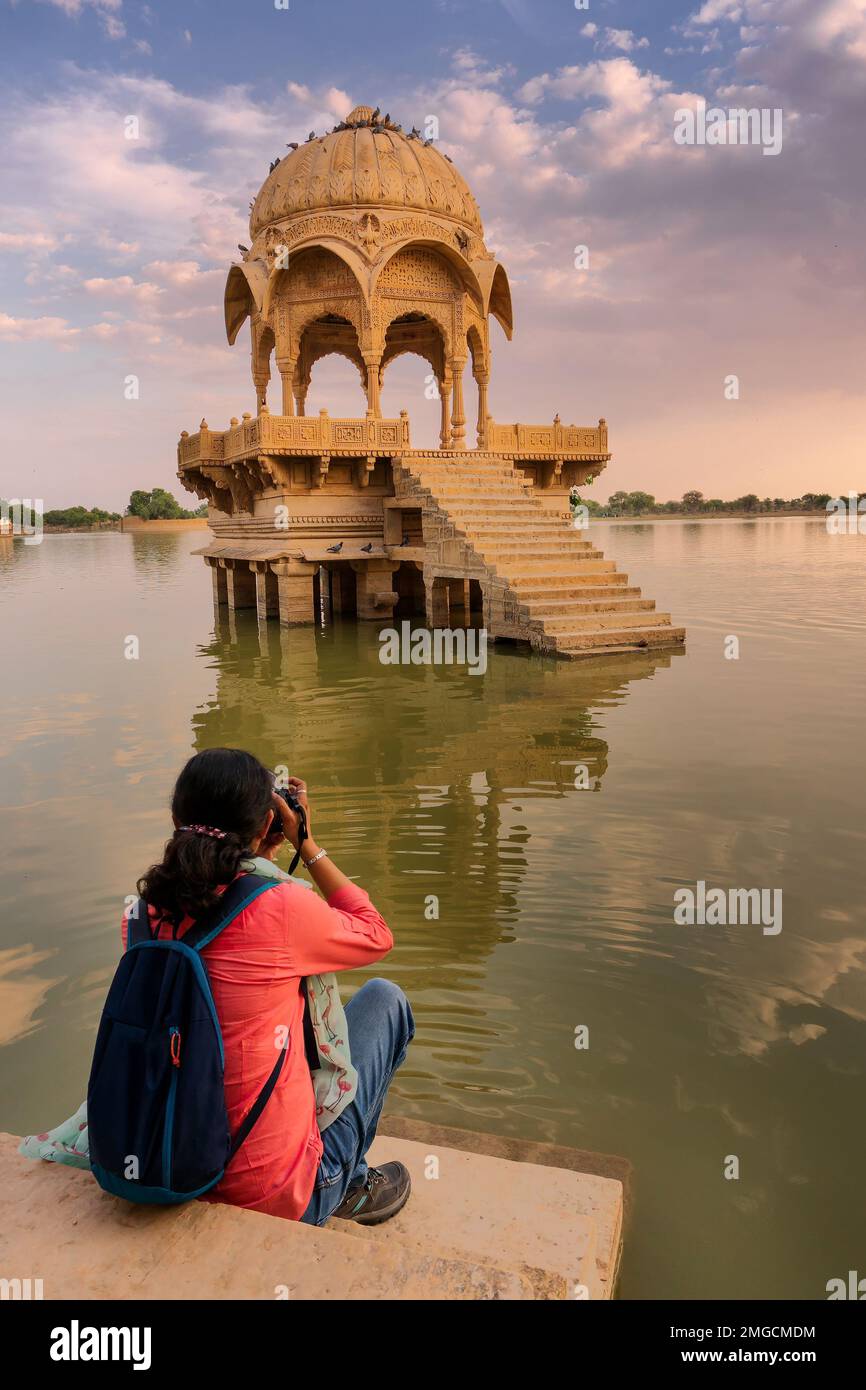 Indian female traveller, woman photographer taking picture of Chhatris and shrines with reflection of them on the water of Gadisar lake. Sun set and c Stock Photo