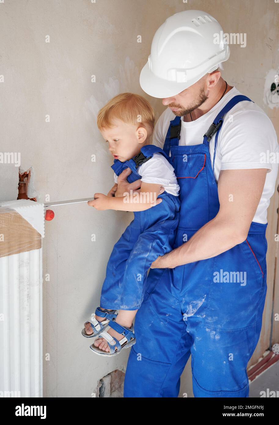 https://c8.alamy.com/comp/2MGFN9J/adorable-child-using-construction-tool-while-installing-heating-radiator-with-father-at-home-male-plumber-in-work-overalls-and-safety-helmet-holding-baby-boy-home-renovation-and-parenting-concept-2MGFN9J.jpg