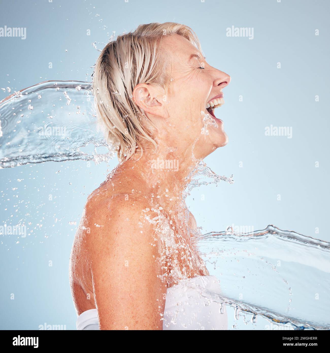Woman, studio and happy with water splash, smile and cleaning for cosmetics by blue background. Excited senior model, skincare shower and cosmetic Stock Photo