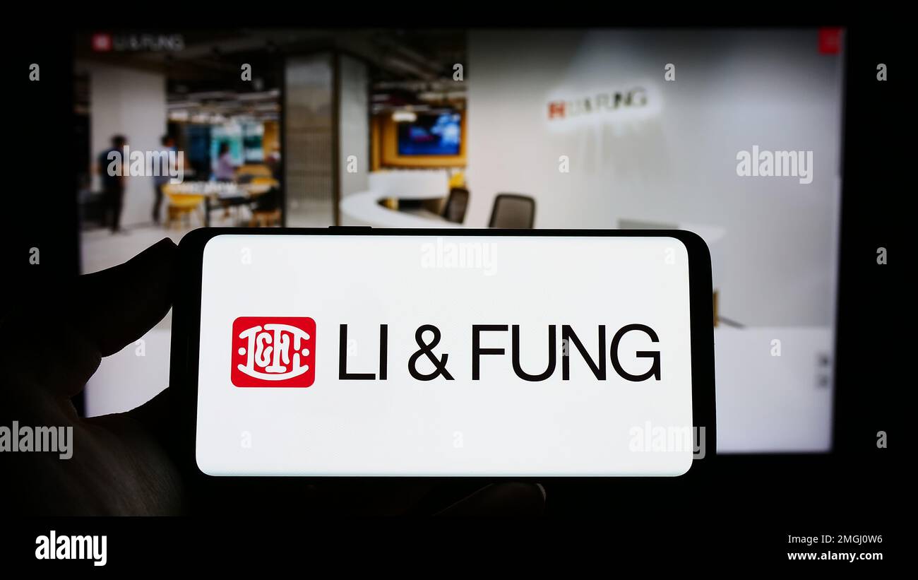 Person holding cellphone with logo of supply chain management company Li Fung Limited on screen in front of webpage. Focus on phone display. Stock Photo