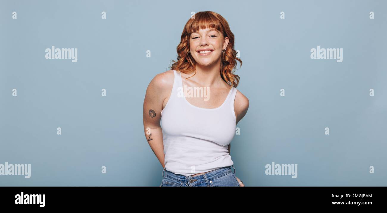 Woman with ginger hair smiling at the camera while wearing a tank top and jeans. Portrait of a happy young woman standing against a blue background. Stock Photo