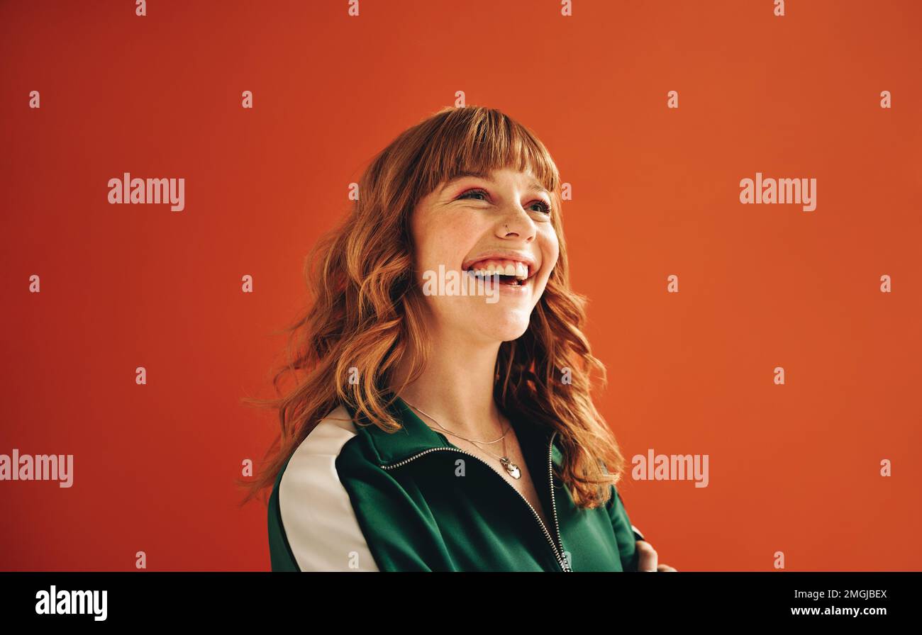 Happy young woman looking away with a smile while standing in a studio. Cheerful woman with ginger hair standing against a vibrant orange background i Stock Photo