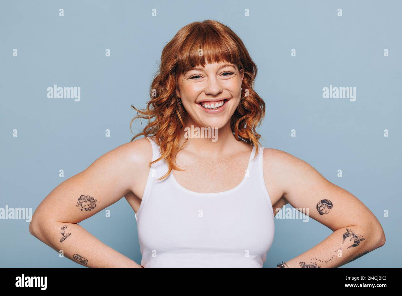 Portrait of a happy young woman with arm tattoos standing in a studio. Cheerful young woman with ginger hair standing against a blue background in a w Stock Photo