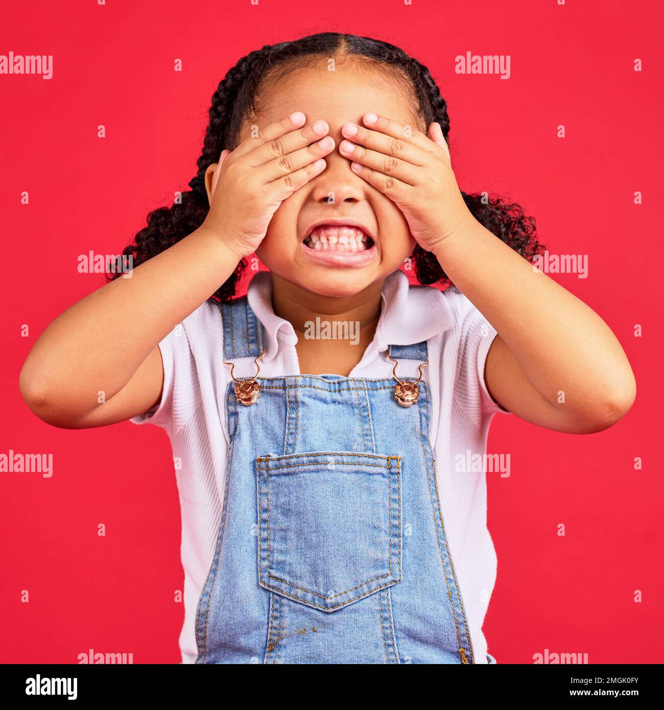 Child, hands and eyes cover on isolated red background with angry, mad or stubborn facial expression. Girl, kid and youth hiding face in tantrum Stock Photo