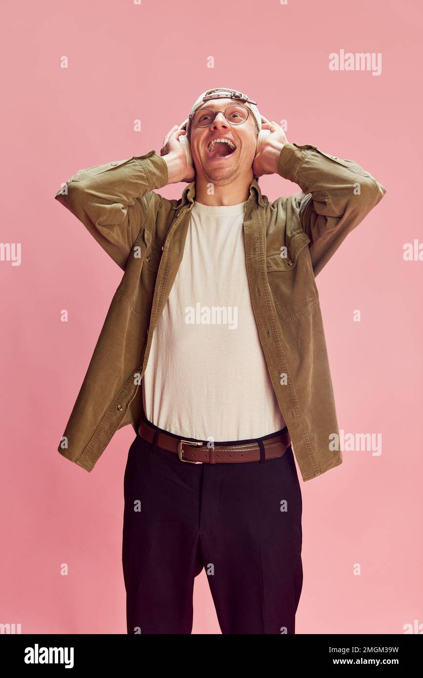 Burst of laugh. Young positive man posing, emotionally listening to music in headphones over pink studio background. Concept of emotions, lifestyle Stock Photo