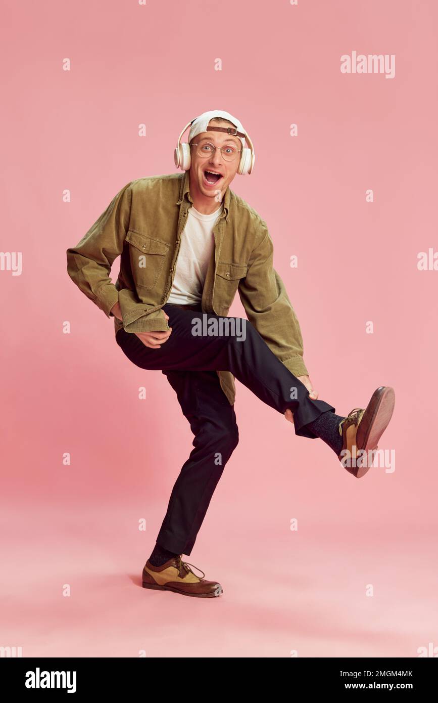 Young positive man cheerfully posing, listening to music in headphones and dancing, having fun over pink studio background. Concept of emotions Stock Photo