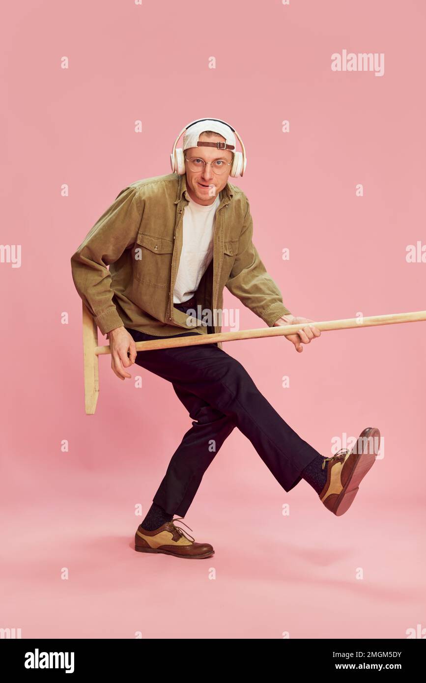 Young emotional man posing, listening to music in headphones, playing with mop like guitar over pink studio background. Concept of emotions, lifestyle Stock Photo