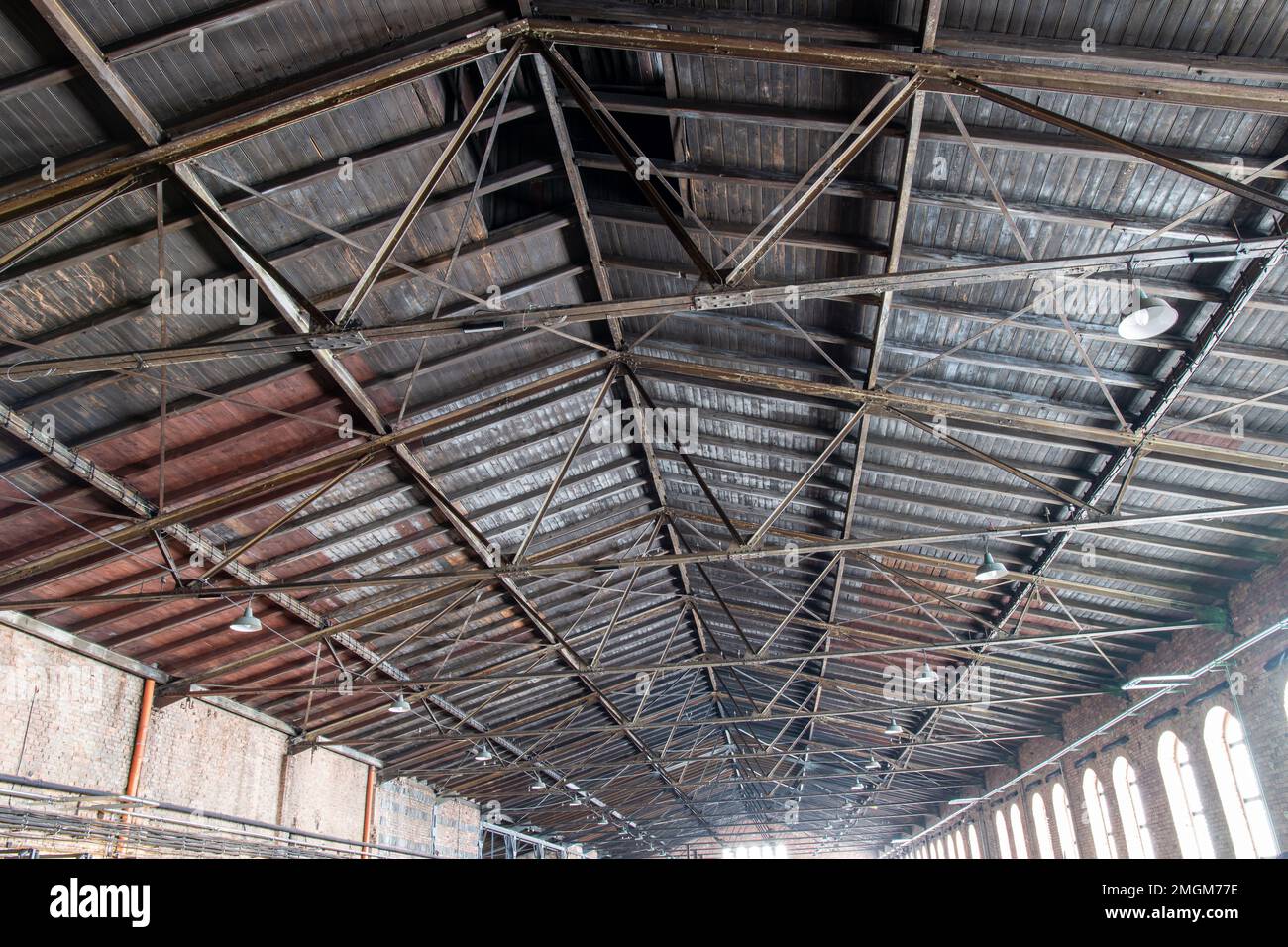 Gable roof truss of a large, vintage factory hall. Roofing construction (sheathing) made of wooden planks. Brick walls and arcade windows. Industrial Stock Photo