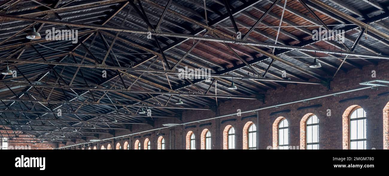 Gable roof truss of a large, vintage factory hall. Roofing construction (sheathing) made of wooden planks. Brick walls and arcade windows. Industrial Stock Photo