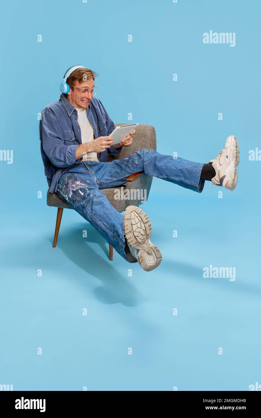 Portrait of youngemotional man in headphones sitting on chair and playing games on tablet over blue studio background. Concept of emotions, lifestyle Stock Photo