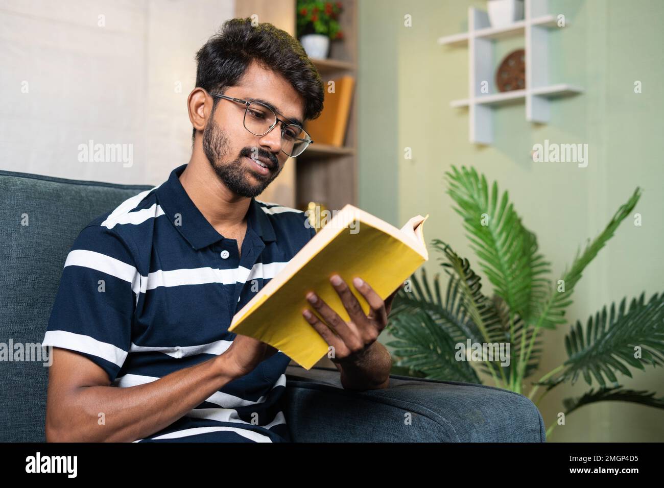 serious young indian student reading book during free time at home - concept of education, hobbies and hard worker. Stock Photo
