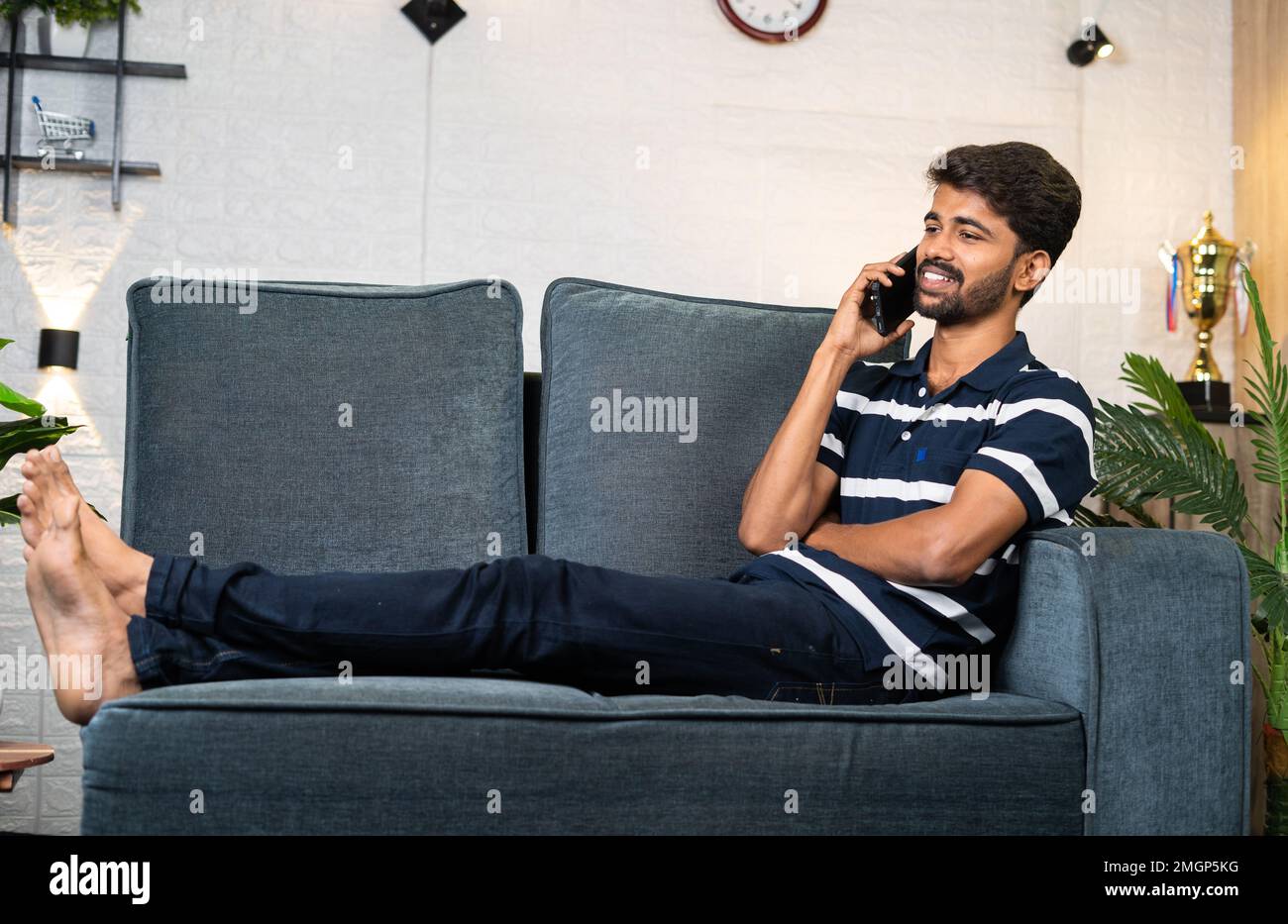 wide shot of Happy smiling young man talking on mobile phone on sofa at home - concept of communication, technology and relaxed conversation. Stock Photo