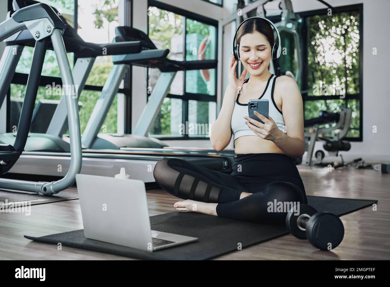 Happy young woman wearing wireless headset and listening to music while resting, sitting on fitness mat after exercising in gym indoors. Stock Photo