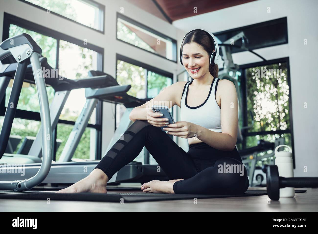 Happy young woman wearing wireless headset and listening to music while resting, sitting on fitness mat after exercising in gym indoors. Stock Photo