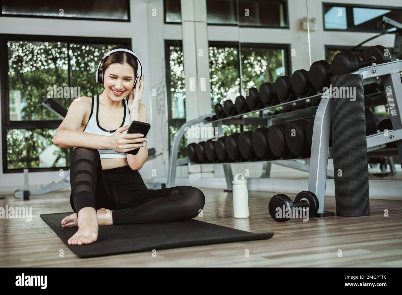 Happy young woman wearing wireless headset and listening to music while resting, sitting on fitness mat after exercising in gym indoors. Stock Photo