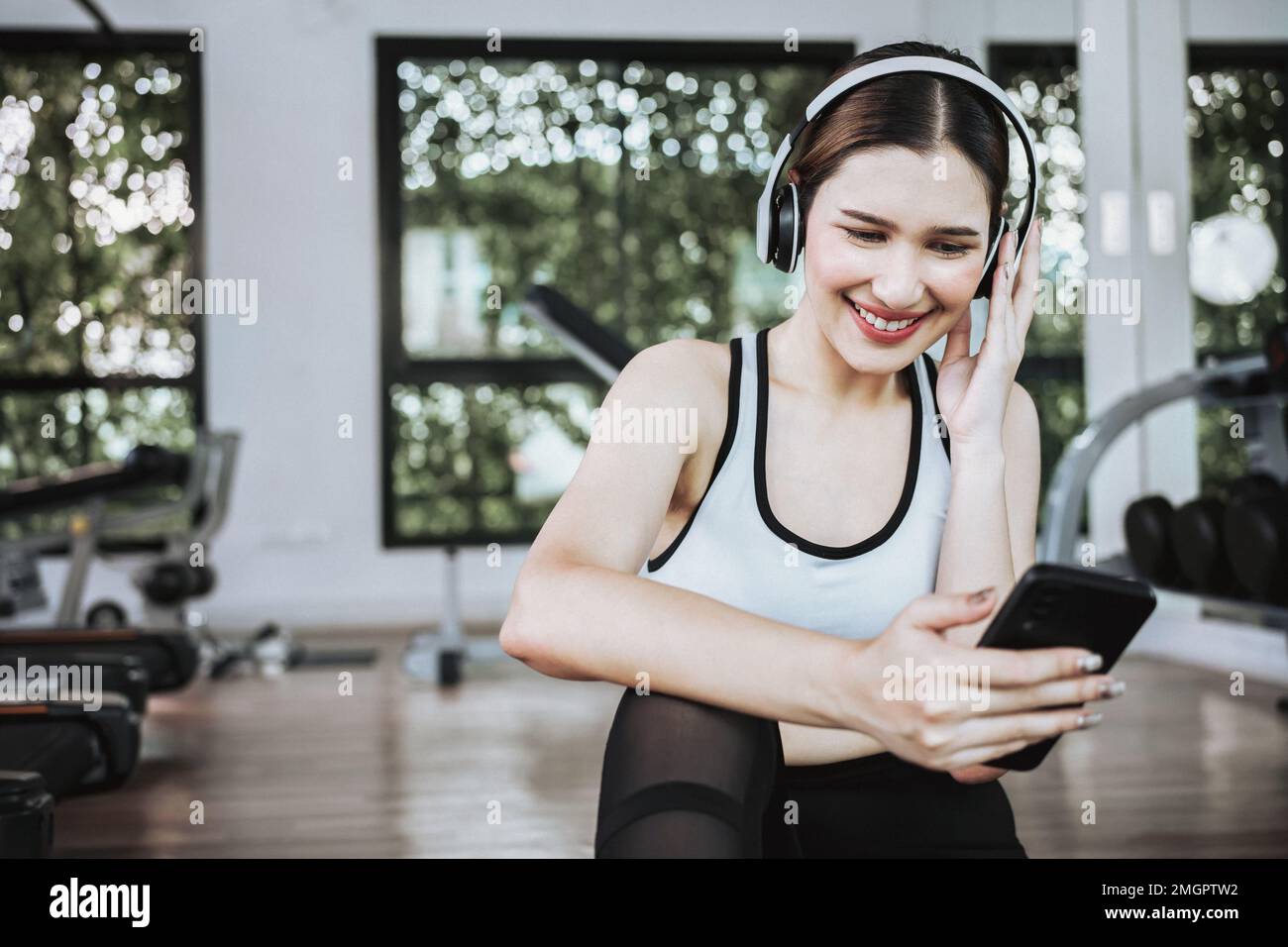 Happy young woman wearing wireless headset and listening to music while resting, sitting on fitness mat after exercising in gym indoors. Stock Photo