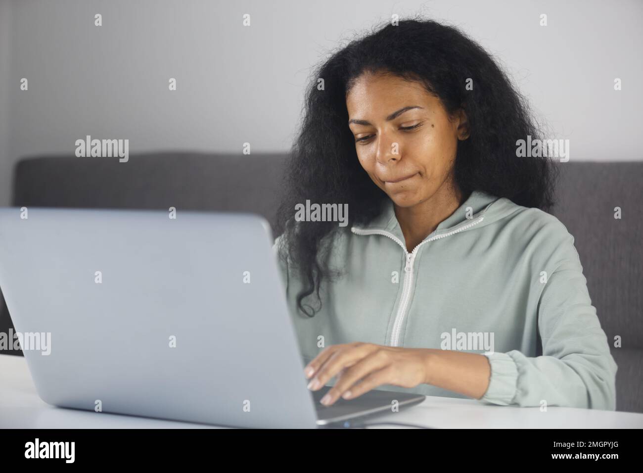Focused black woman biting lip while typing text message on laptop keyboard. BIPOC entrepreneur female working on notebook computer at home Stock Photo