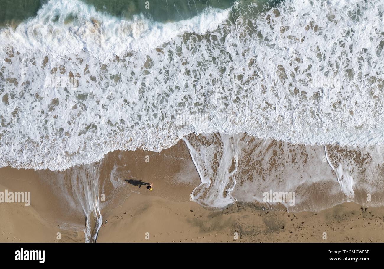 Aerial drone point of view of person walking on sand in a beach. Stormy waves idyllic beach in winter. Stock Photo