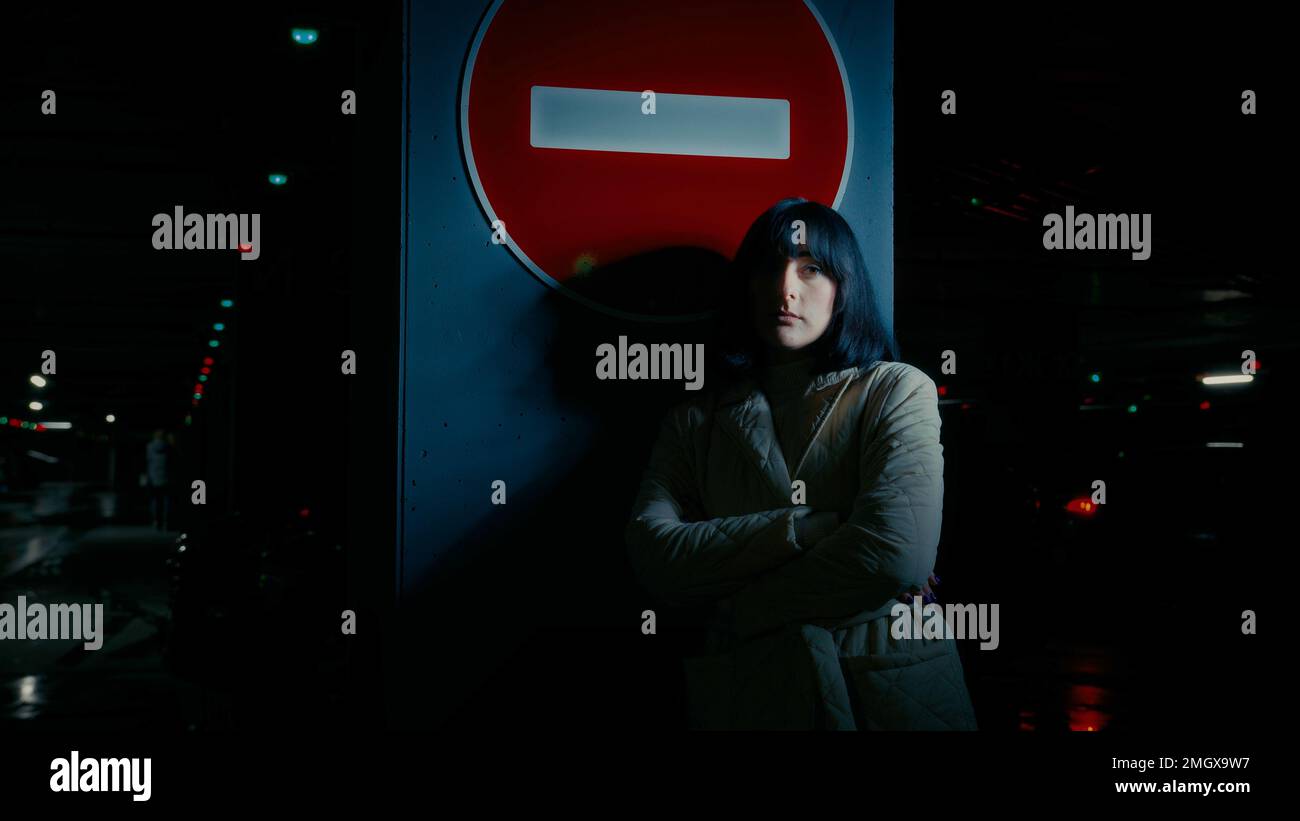 Sad upset stressed anxious depressed woman alone girl female lady standing at dark parking lot at night darkness near stop traffic road sign no drive Stock Photo