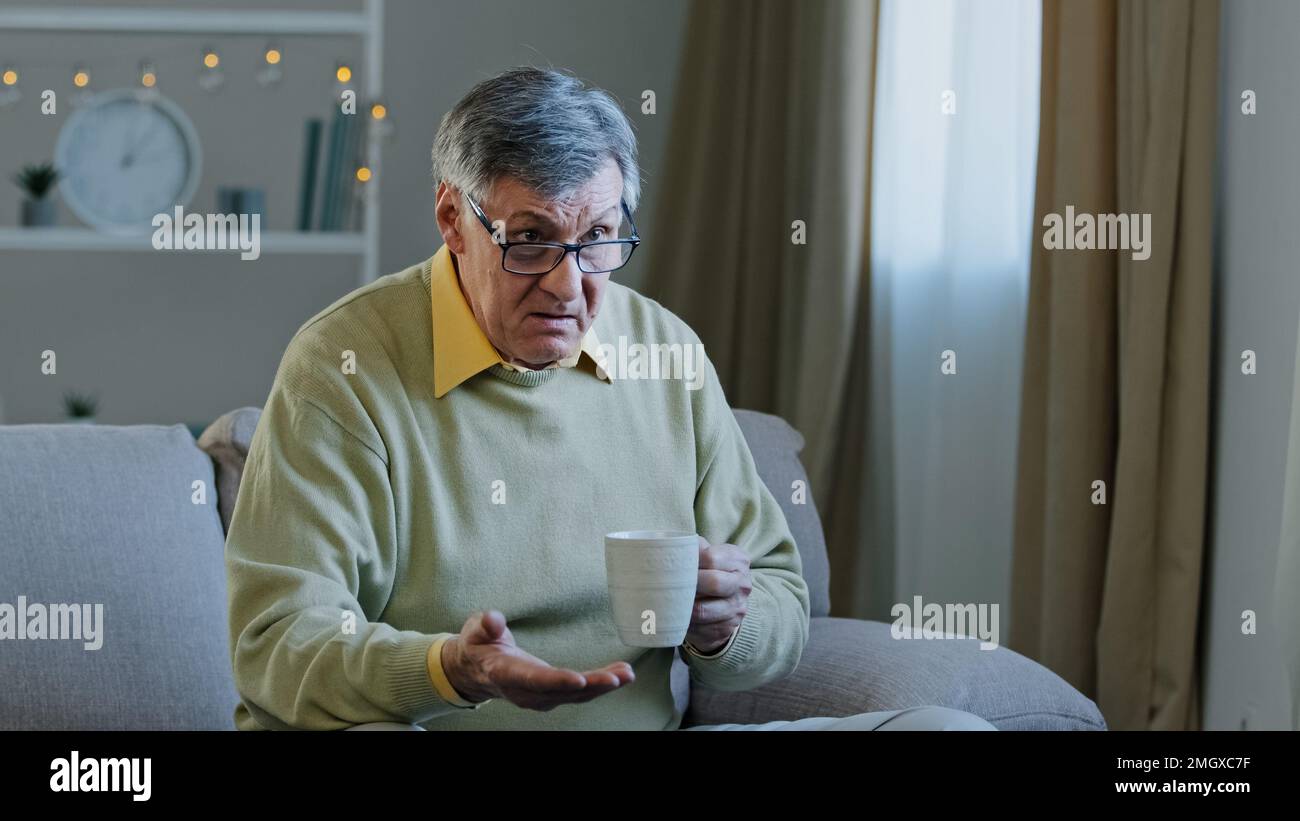 Old elderly mature grandfather man sitting at home couch watching TV drinking hot tea from cup supporting team in online sports competition frustrated Stock Photo