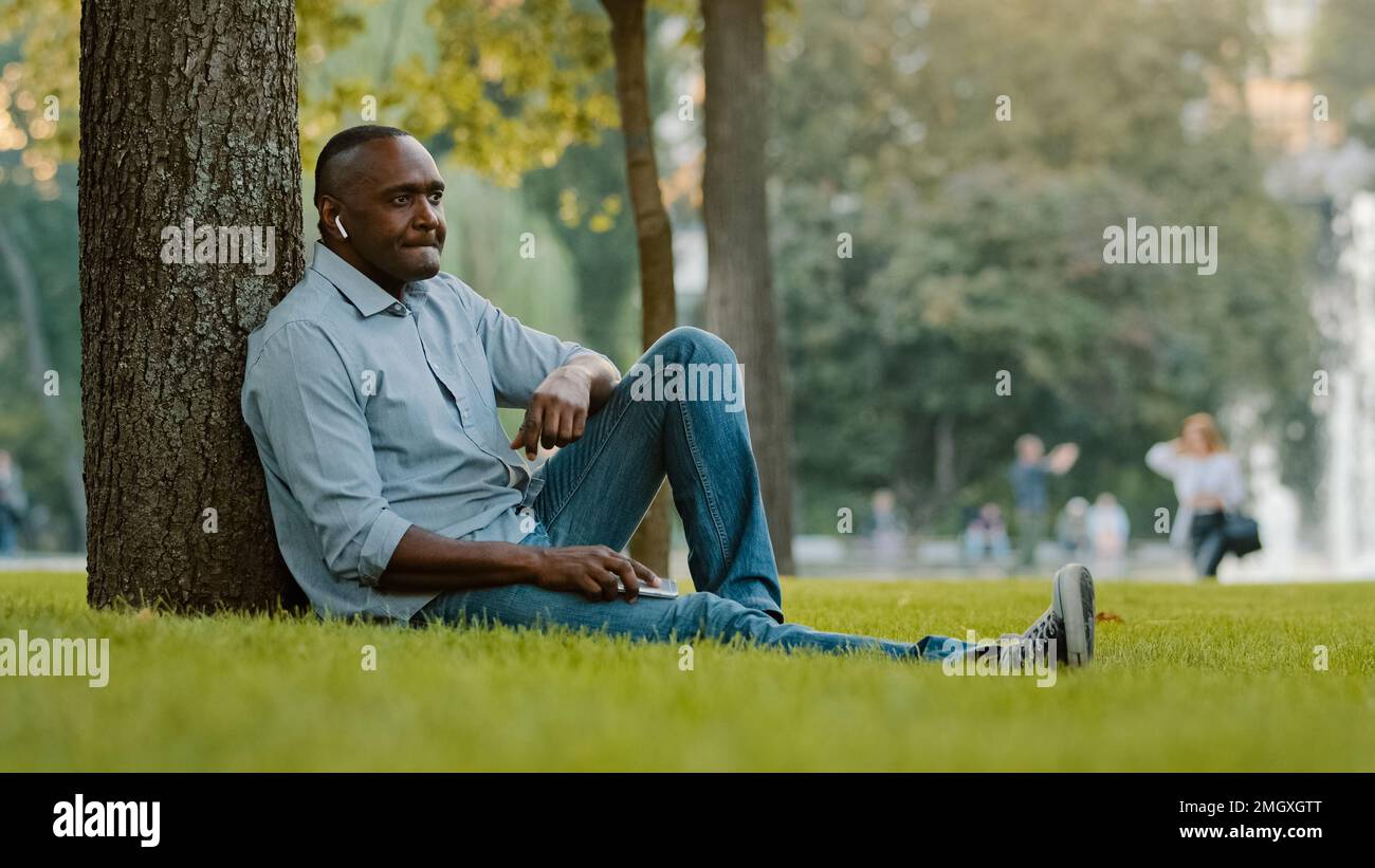 Calm mature adult male enjoying weekend outdoors listening song in wireless headphones with great sound. Senior middle-aged African American man Stock Photo
