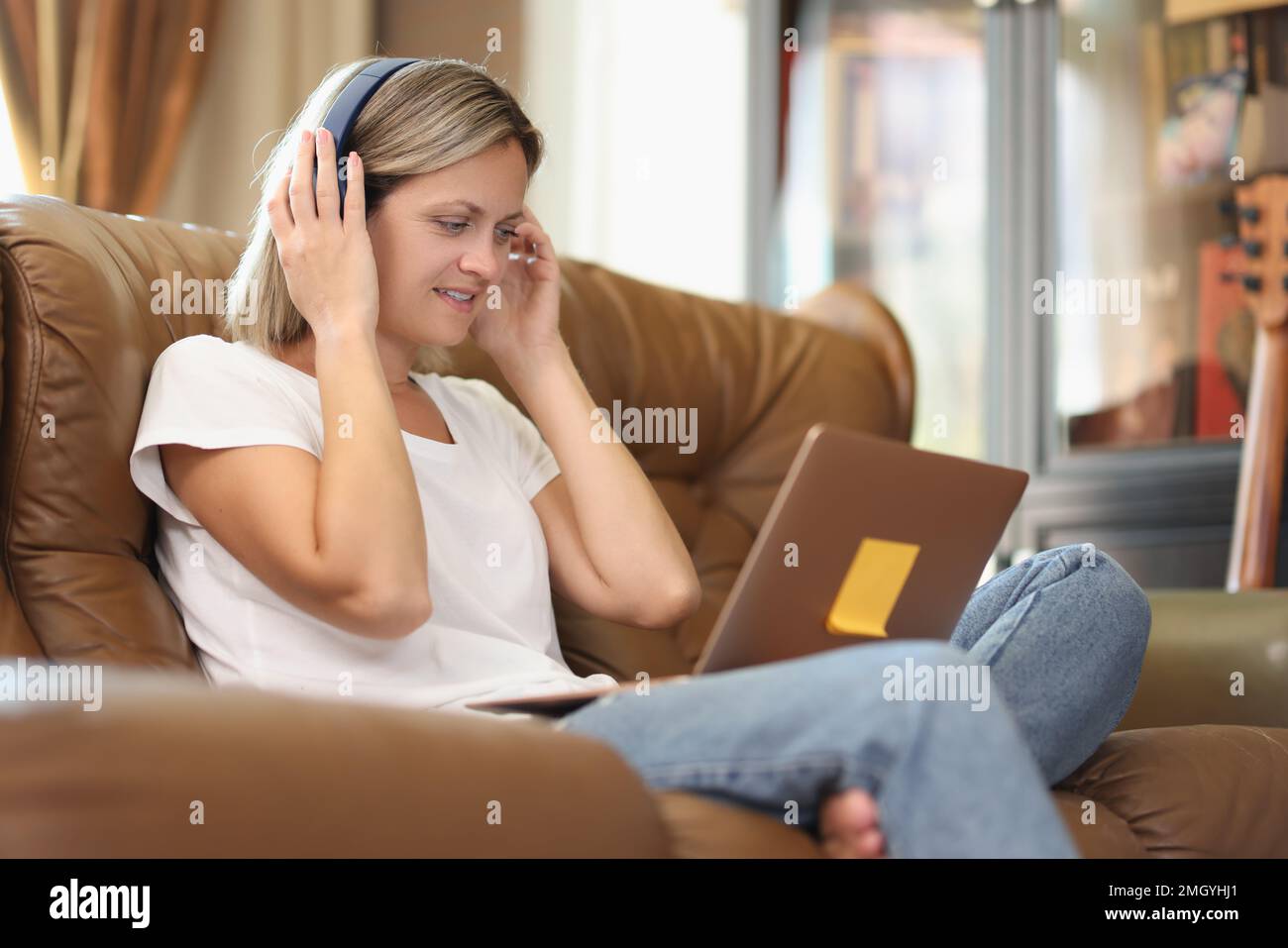 Smiling woman with wireless headphones and laptop sitting on sofa at home. Stock Photo