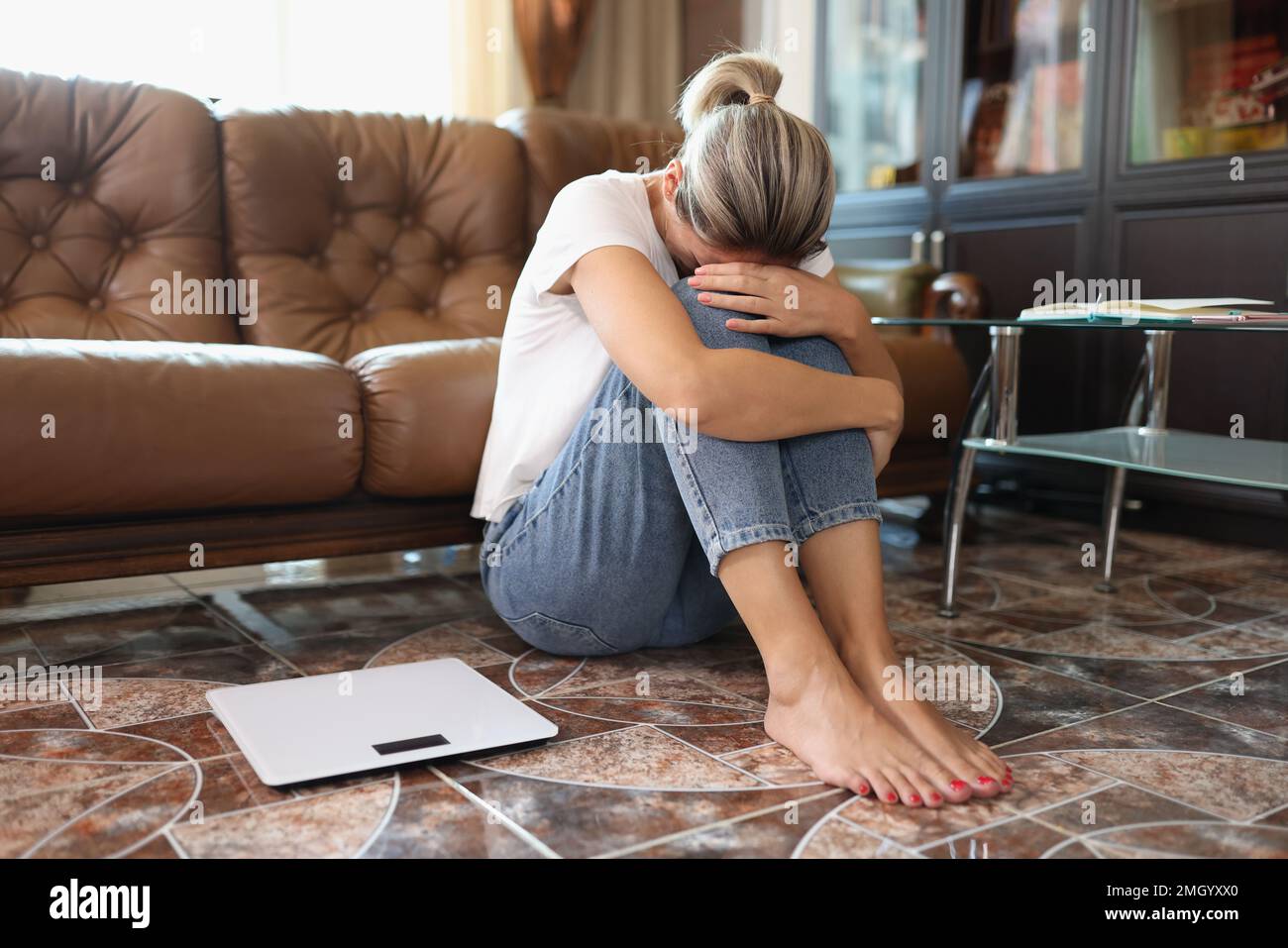 Sad woman sits on floor near scales and holding her knees by hands. Stock Photo