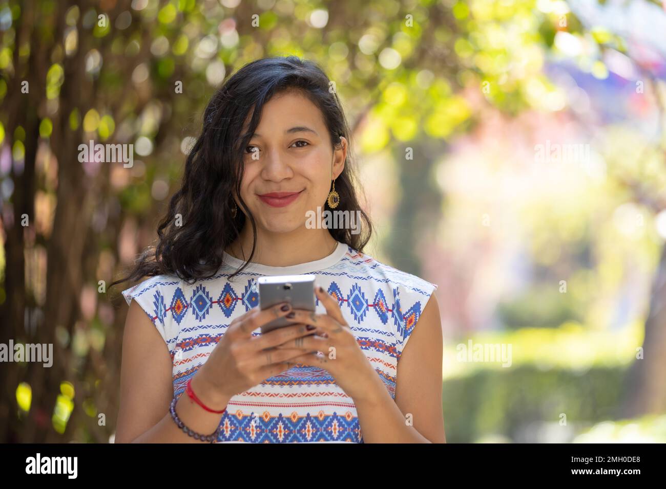 Real Mexican woman holding smart phone and looking at camera outdoors Stock Photo
